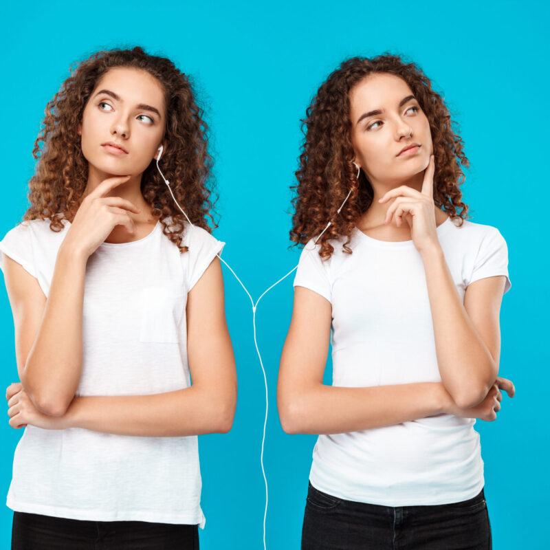 Two young pretty girls twins listening music in headphones, thinking over blue background. Copy space.